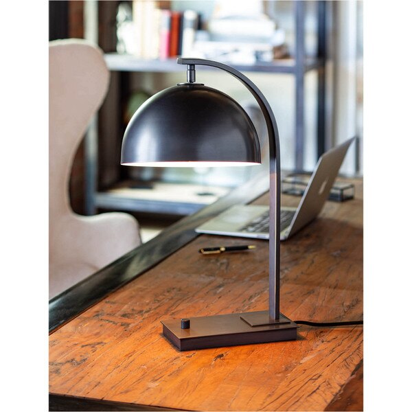 adjustable metal desk lamp on wooden desk with bookshelf and laptop in background